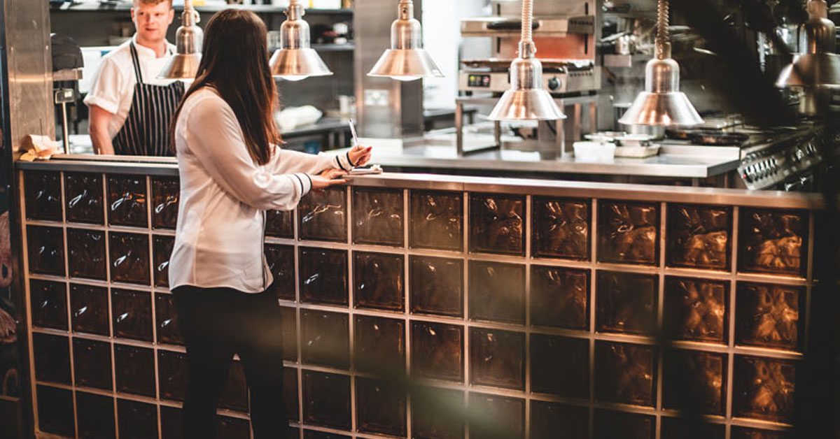 Waitress at bar wearing black, slip-resistant Radium shoes with restaurant kitchen in background
