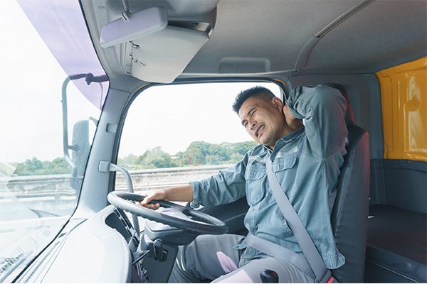 HGV driver with strained expression sitting with one hand on the wheel and rubbing his neck with the other