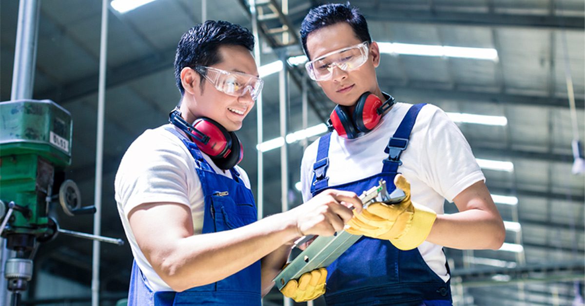 Two manufacturing workers inspecting a metal sheet while wearing safety glasses and noise-cancelling headphones