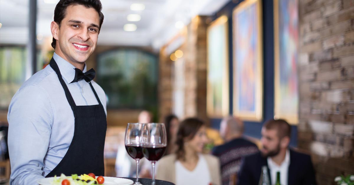 Un camarero sonríe a la cámara mientras lleva una bandeja con un plato y dos copas de vino
