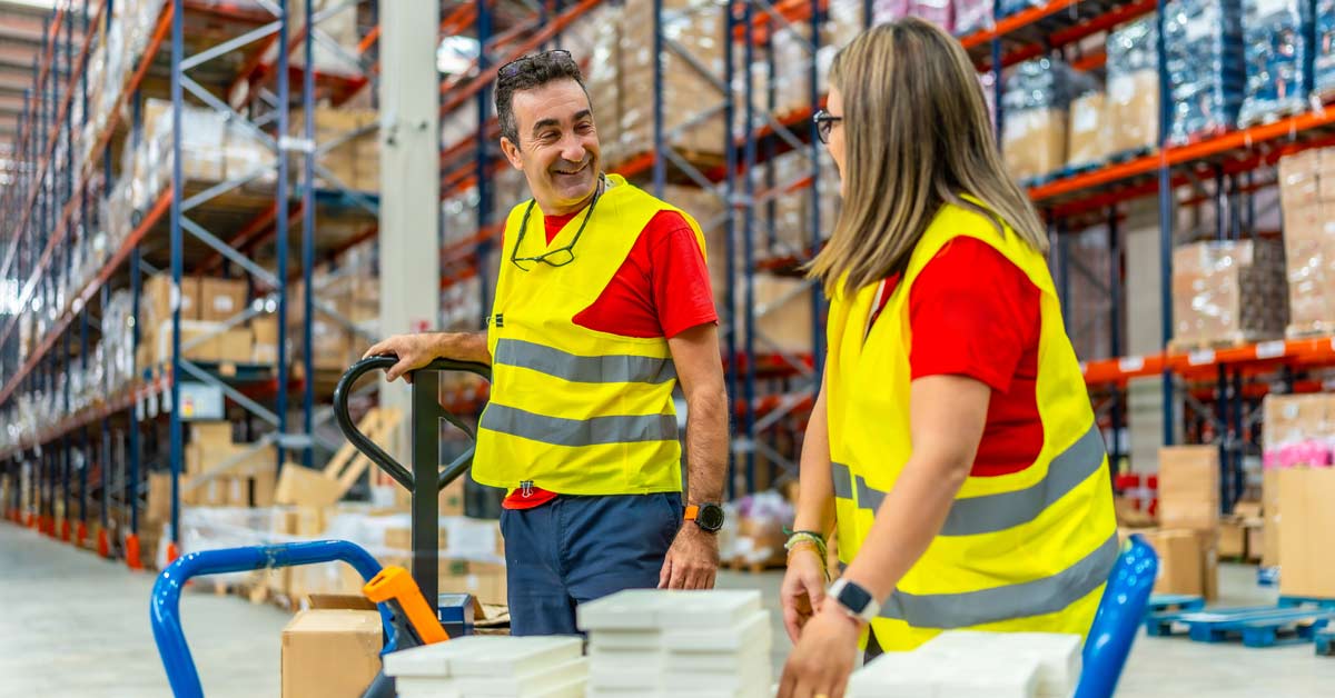 Dos trabajadores de almacén conversan y sonríen mientras trasladan los productos