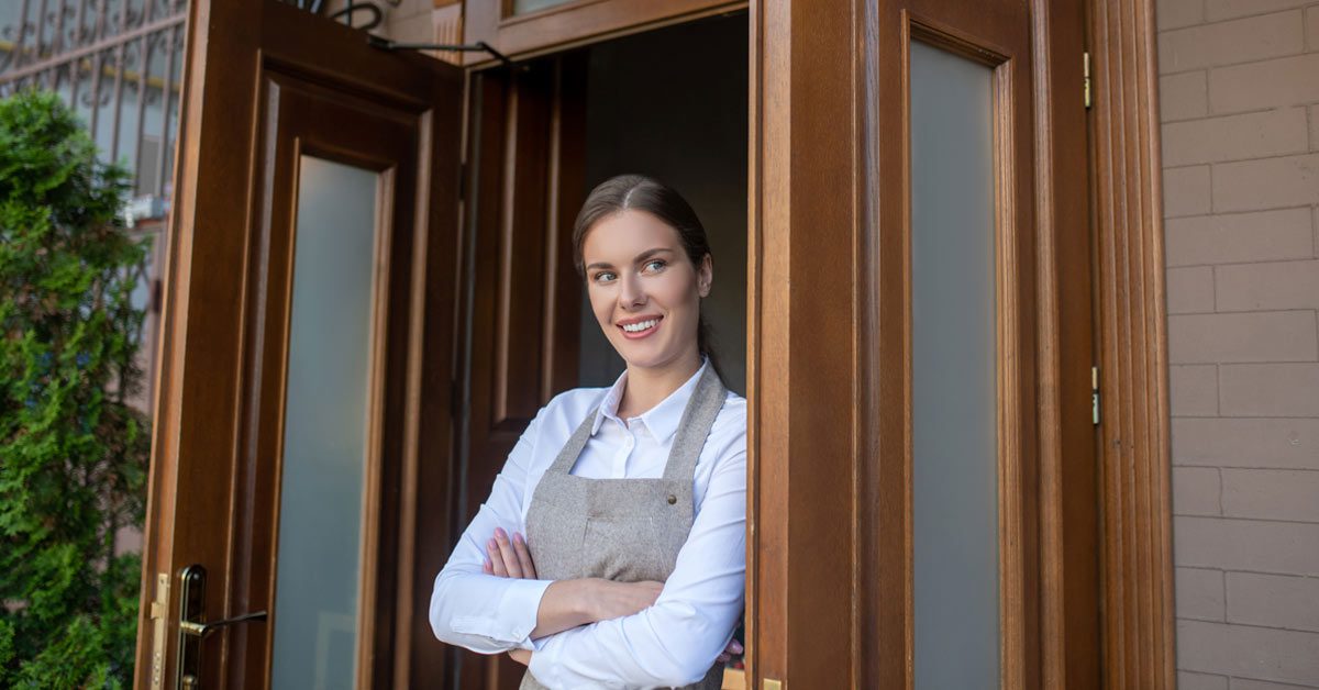 Una joven con delantal sonríe en la puerta de un establecimiento de hostelería